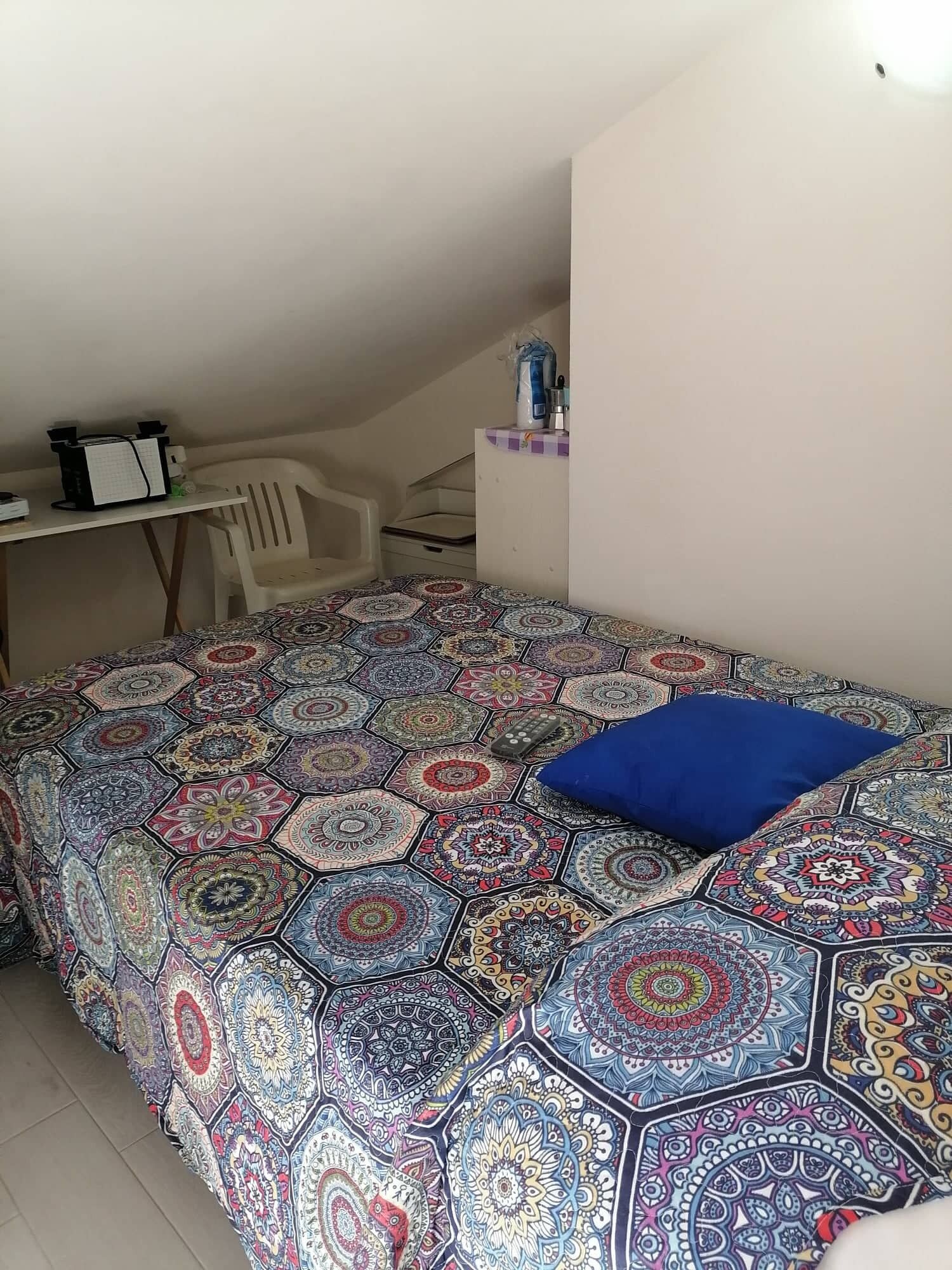 A bedroom with a colorful patterned bedspread, a blue pillow, a desk with a computer and a chair, and a small shelf with items.