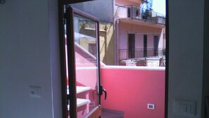 On aperçoit, à travers une porte vitrée, une cour intérieure avec un mur rouge, un banc en bois et un escalier menant à un étage supérieur.