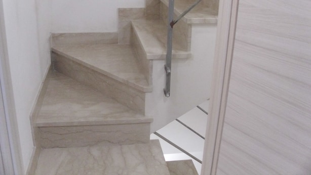 Un escalier étroit doté d’une main courante en métal, menant à un palier avec une rambarde blanche.