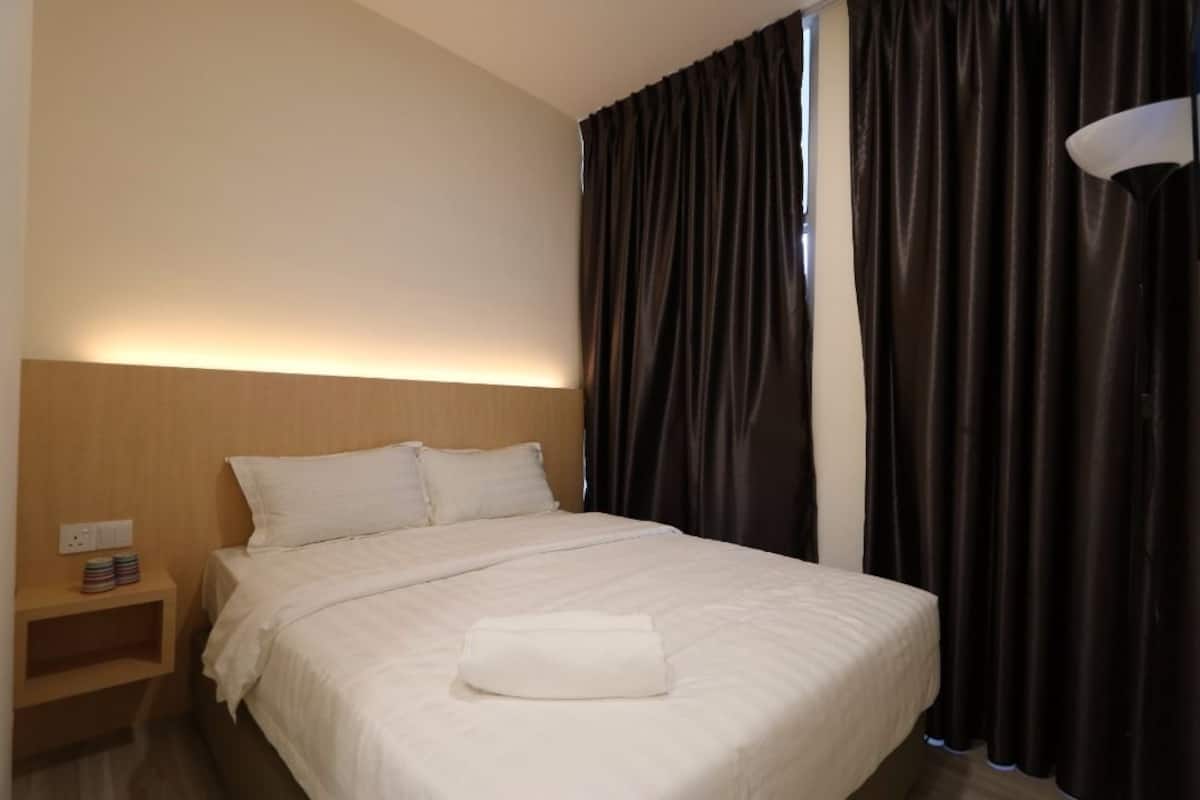 A neatly made bed with white linens, a bedside table with a lamp, and dark curtains.