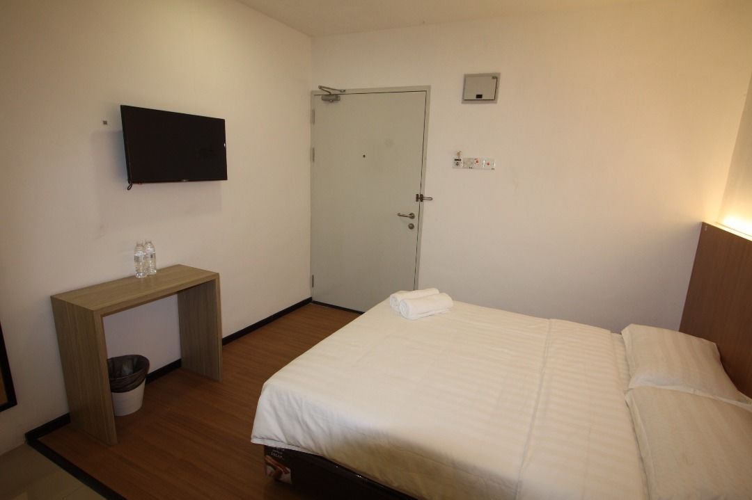 Habitación de hotel con cama, escritorio, silla, televisor, puerta y un pequeño cubo de basura.