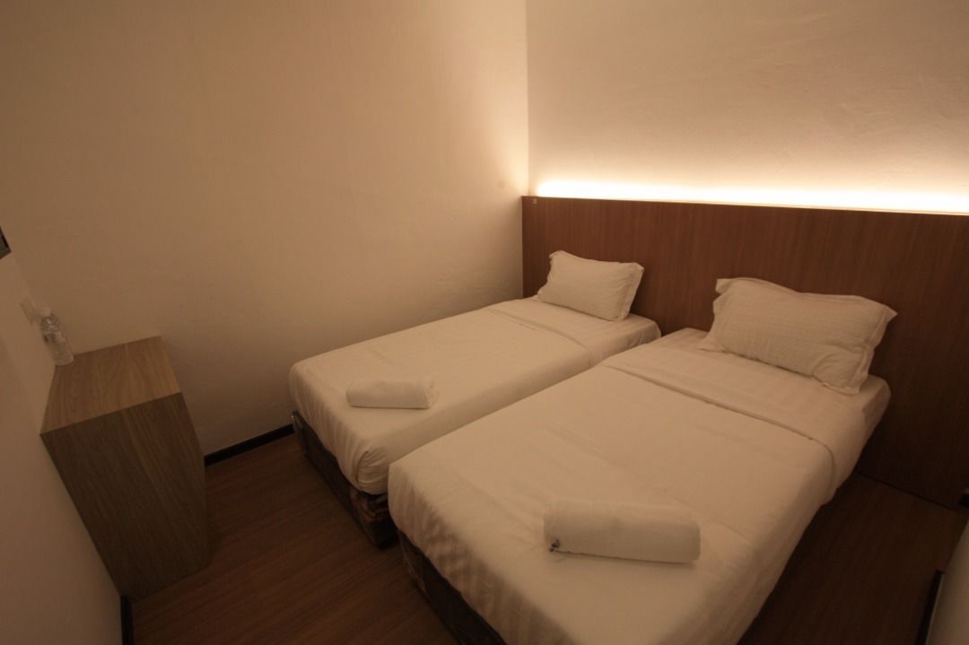 Una habitación pequeña y limpia con dos camas individuales, una mesita de noche de madera y una lámpara fijada a la pared.