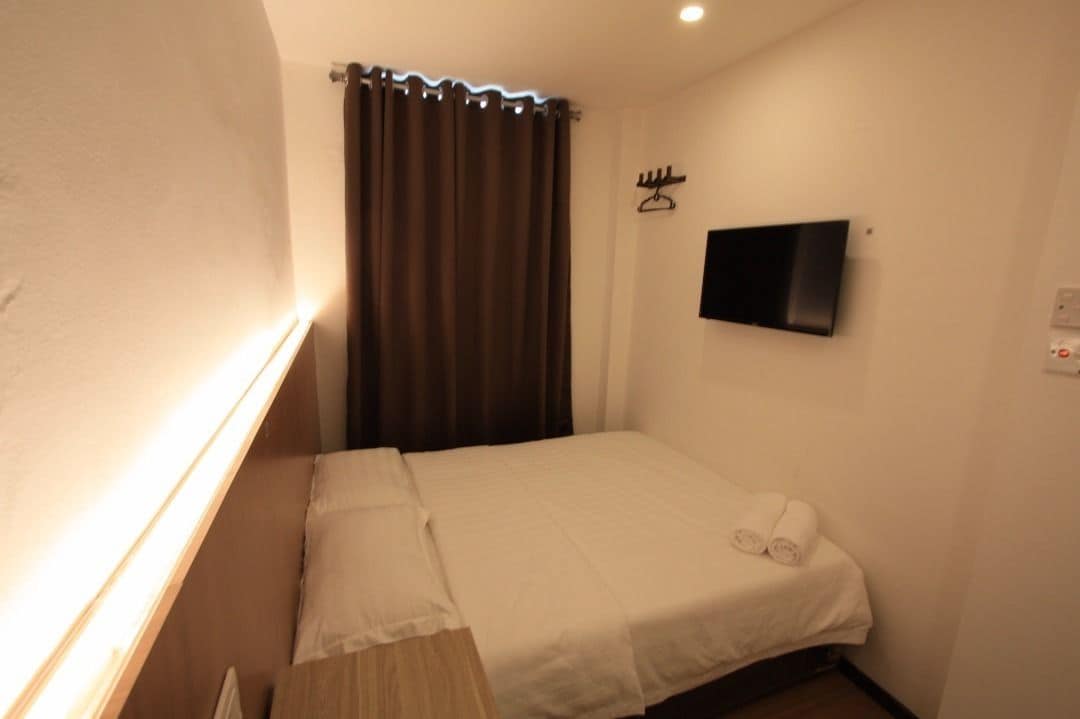 Un petit lit bien fait, avec des draps blancs et une serviette enroulée, un téléviseur à écran plat fixé au mur et un rideau tiré à moitié sur le côté.