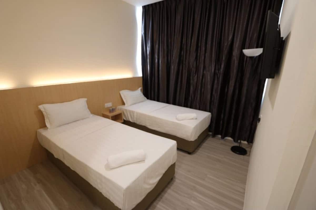 A hotel room with two single beds, a wooden headboard, a small bedside table, a wall-mounted TV, and a large window with dark curtains.