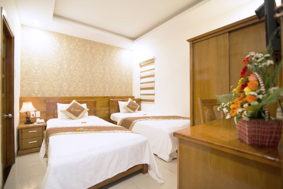 A hotel room with two beds, a wooden headboard, a nightstand with a lamp, and a wooden dresser with a vase of flowers.