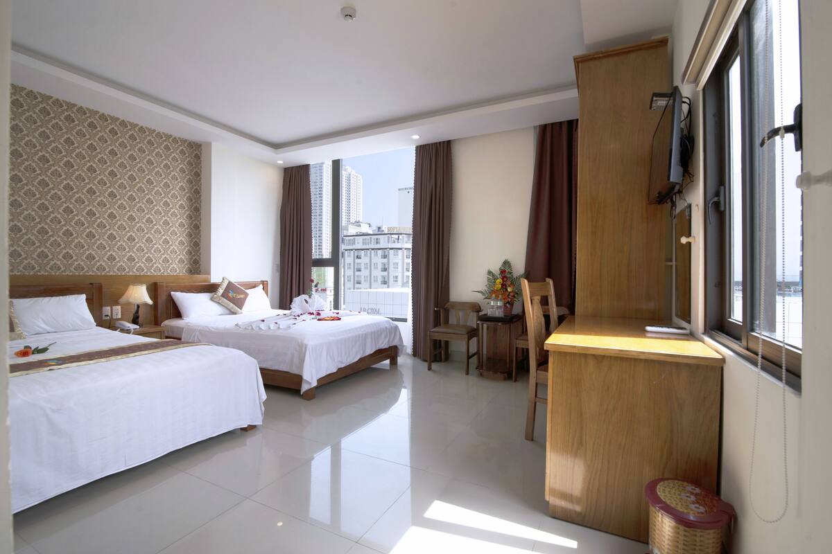 A hotel room with two beds, a desk, a chair, and a view of the city.