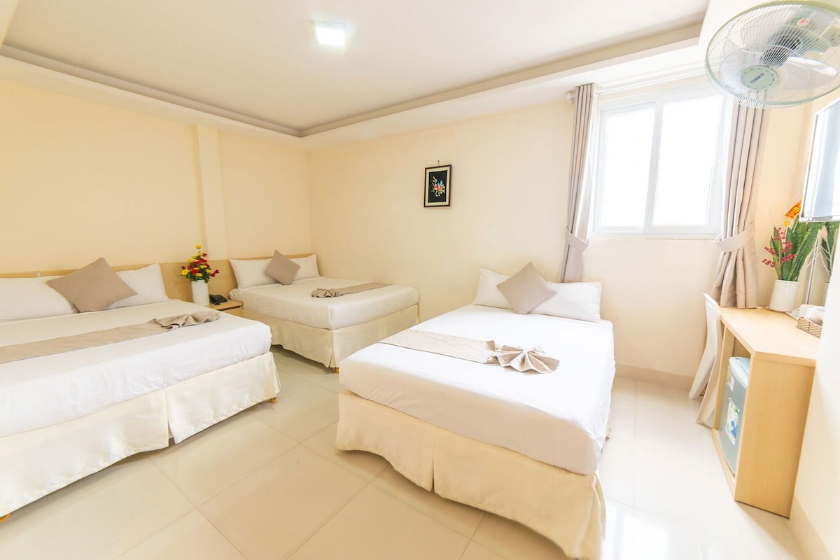 A hotel room with two beds, a desk, a chair, a fan, and a window with curtains.