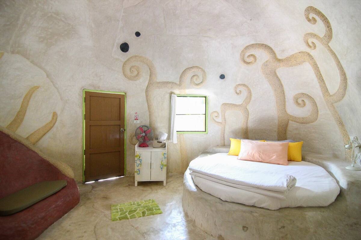 A bedroom with a bed, a door, a fan, and a small cabinet.