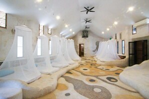 A room with multiple beds covered in white canopies, a patterned carpet, and a ceiling fan. - Naidee Sculptured Huts (Hua Hin)