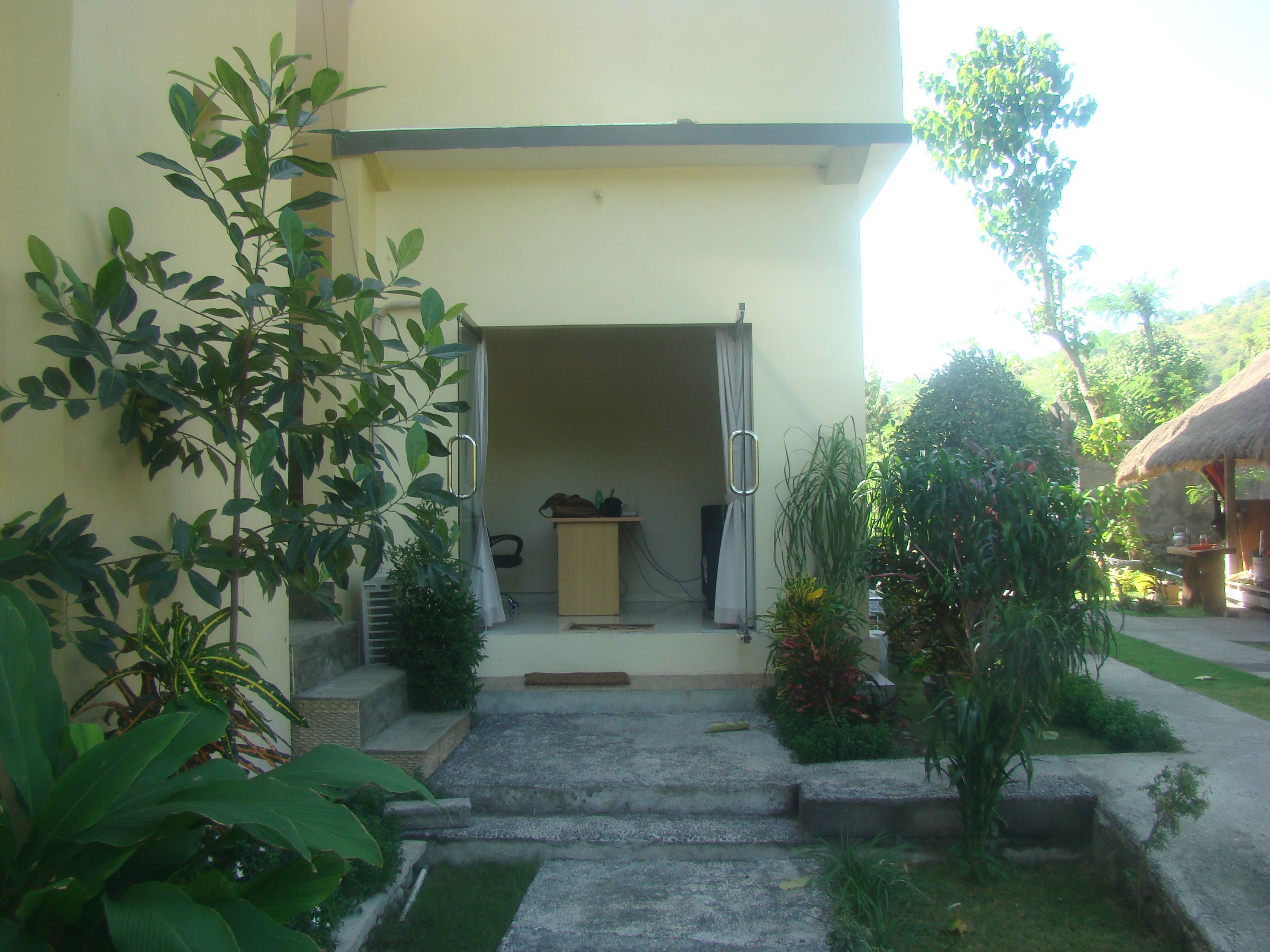 A building entrance with a concrete pathway leading to a door, surrounded by various plants and a small structure with a thatched roof.