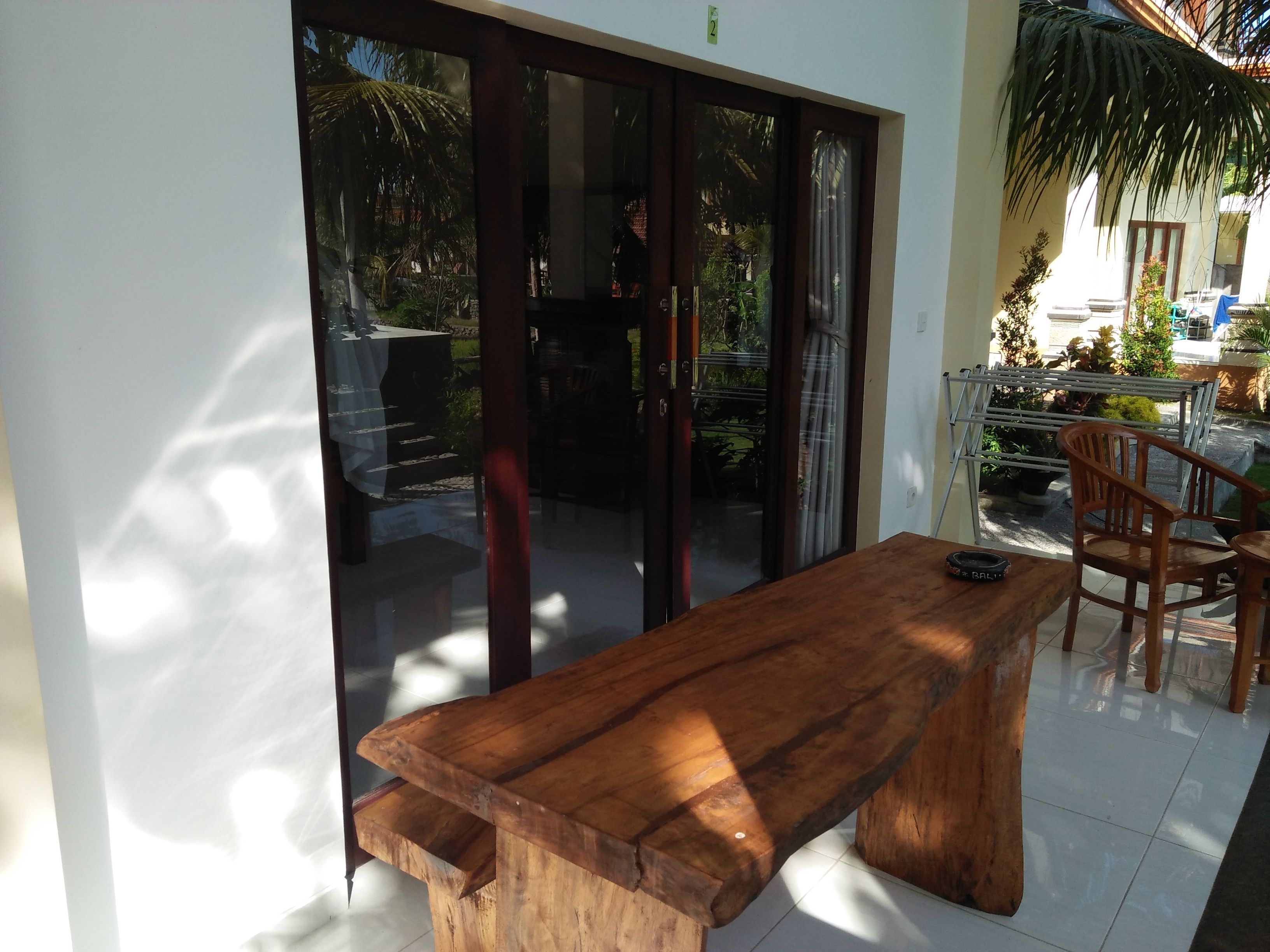 Una mesa y sillas de madera en una habitación iluminada por el sol, con un ventanal corredizo que da a un espacio exterior con plantas y vistas al cielo.