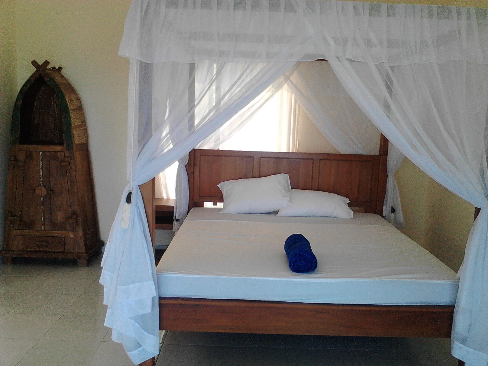 Uma cama com cabeceira de madeira e roupa de cama branca, uma toalha azul enrolada e um armário de madeira à esquerda.