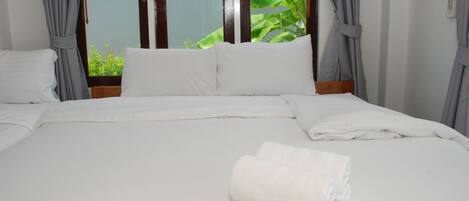 Una cama bien hecha con sábanas blancas y una toalla doblada, cortinas y una ventana con vistas a la vegetación.