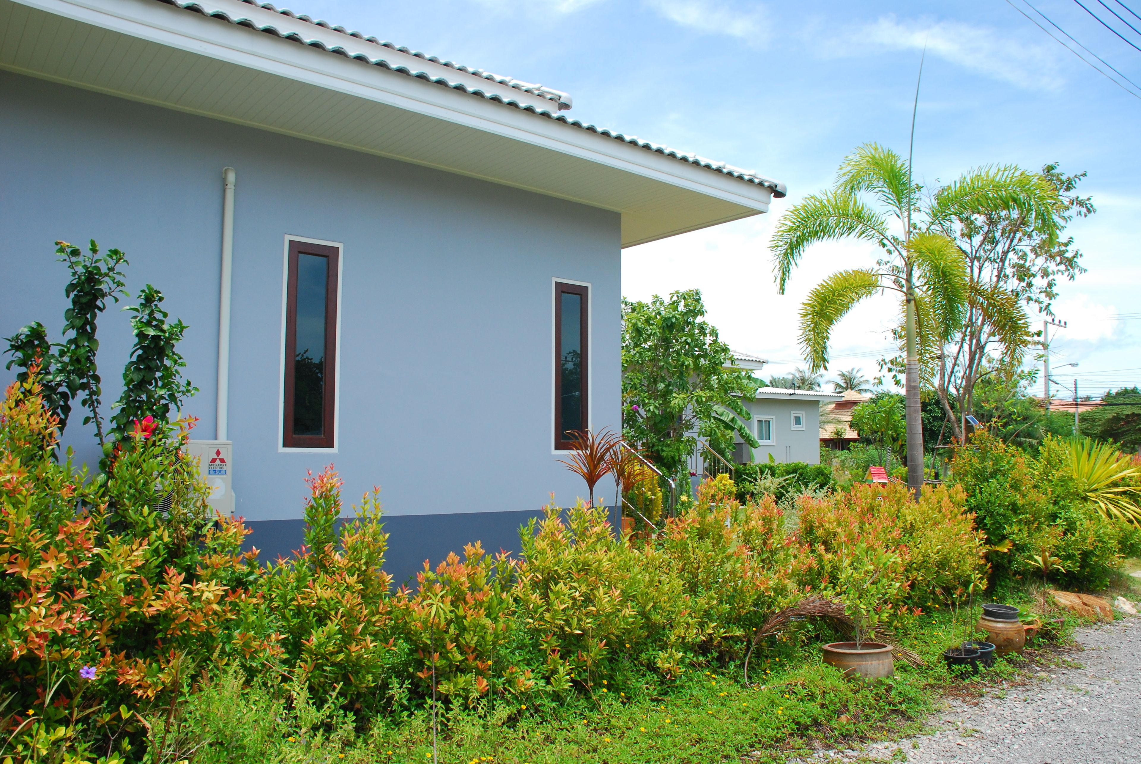A two-story house with a light blue exterior and a white roof. There are two windows on the front side, and the house is surrounded by various plants and shrubs.