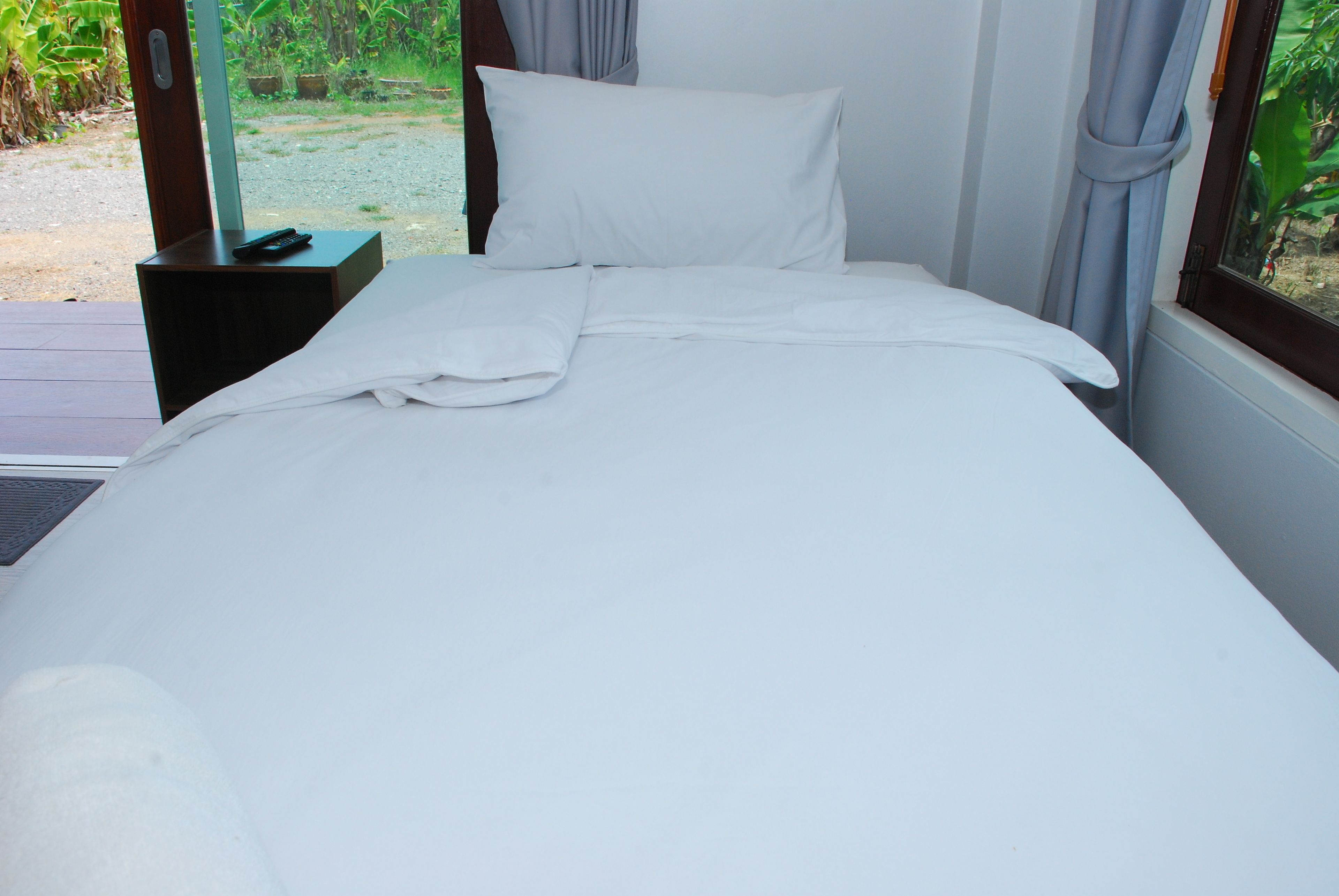 Una cama bien tendida con sábanas blancas, un control remoto en una mesita de noche y vistas al verdor exterior.