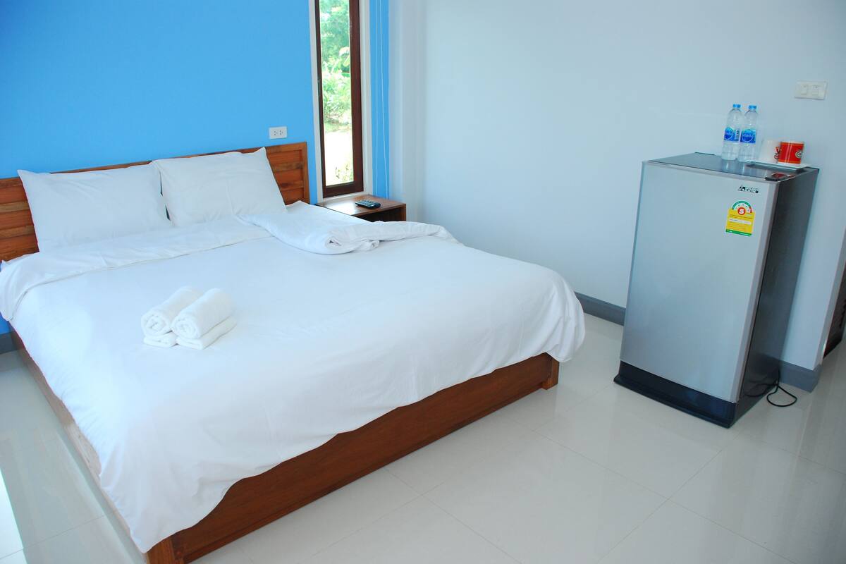 A neatly made bed with white linens and towels, a small bedside table with a remote control, and a refrigerator with water bottles on top.