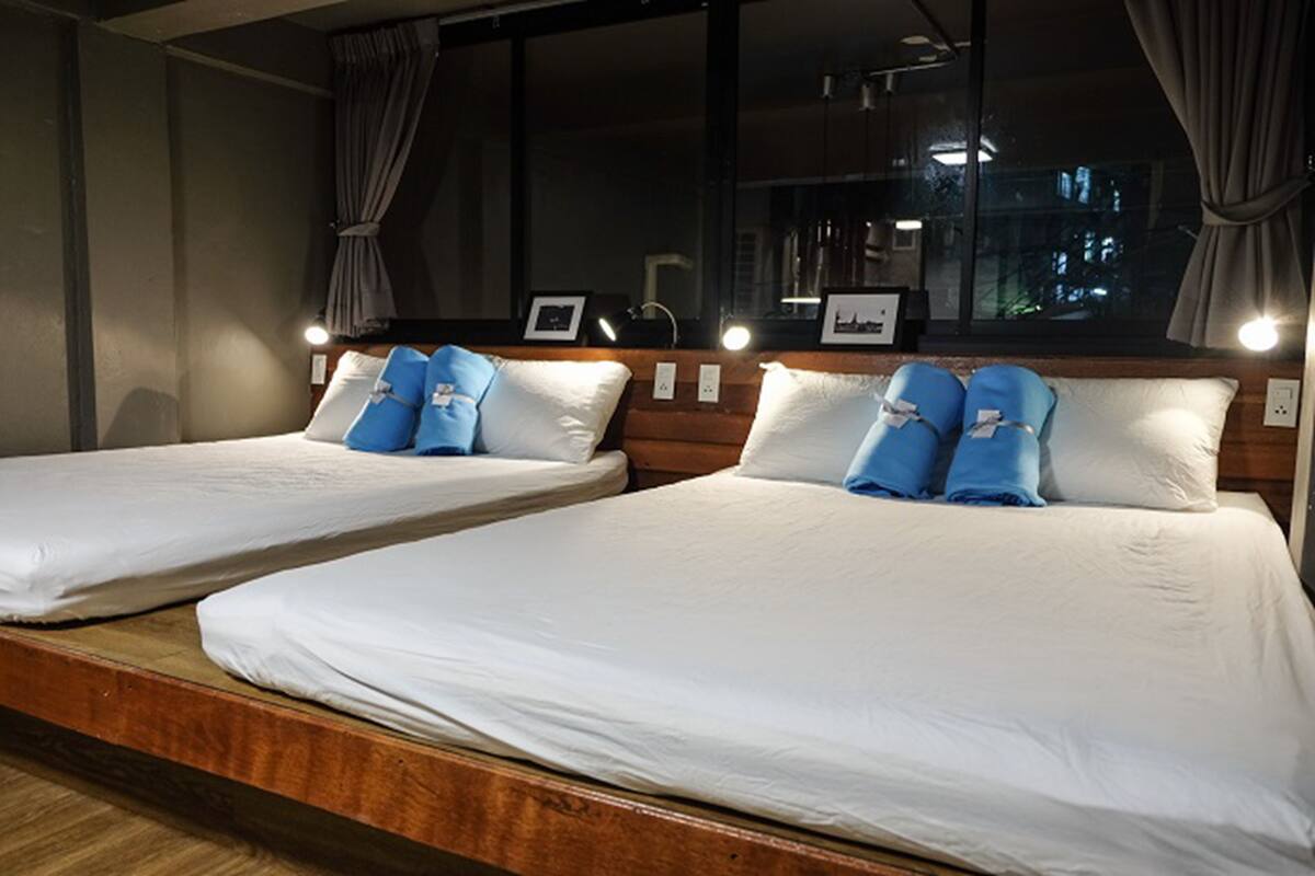 Two single beds with white linens and blue pillows, a wooden headboard, and a large window with curtains.