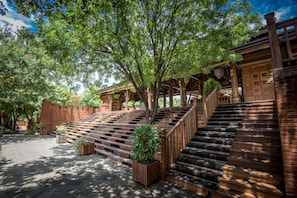 Un escalier en bois mène à un bâtiment doté d’un porche abrité, qui est entouré d’arbres et de plantes en pot.