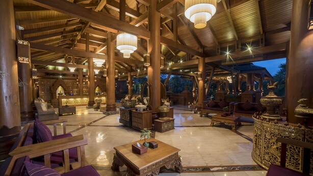 A spacious lobby with high ceilings, wooden beams, and chandeliers. There are seating areas with purple cushions and a reception desk.