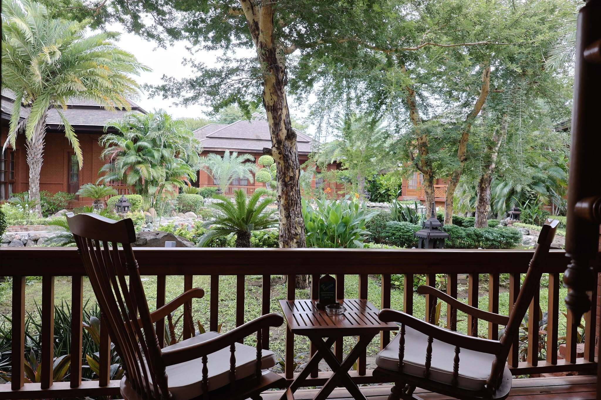 Una terraza de madera con dos sillas y una mesa, con vistas a un jardín exuberante con varias plantas y un edificio al fondo.