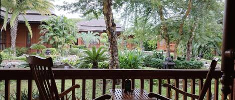 Una terraza de madera con dos sillas y una mesa, con vistas a un jardín exuberante con varias plantas y un edificio al fondo.