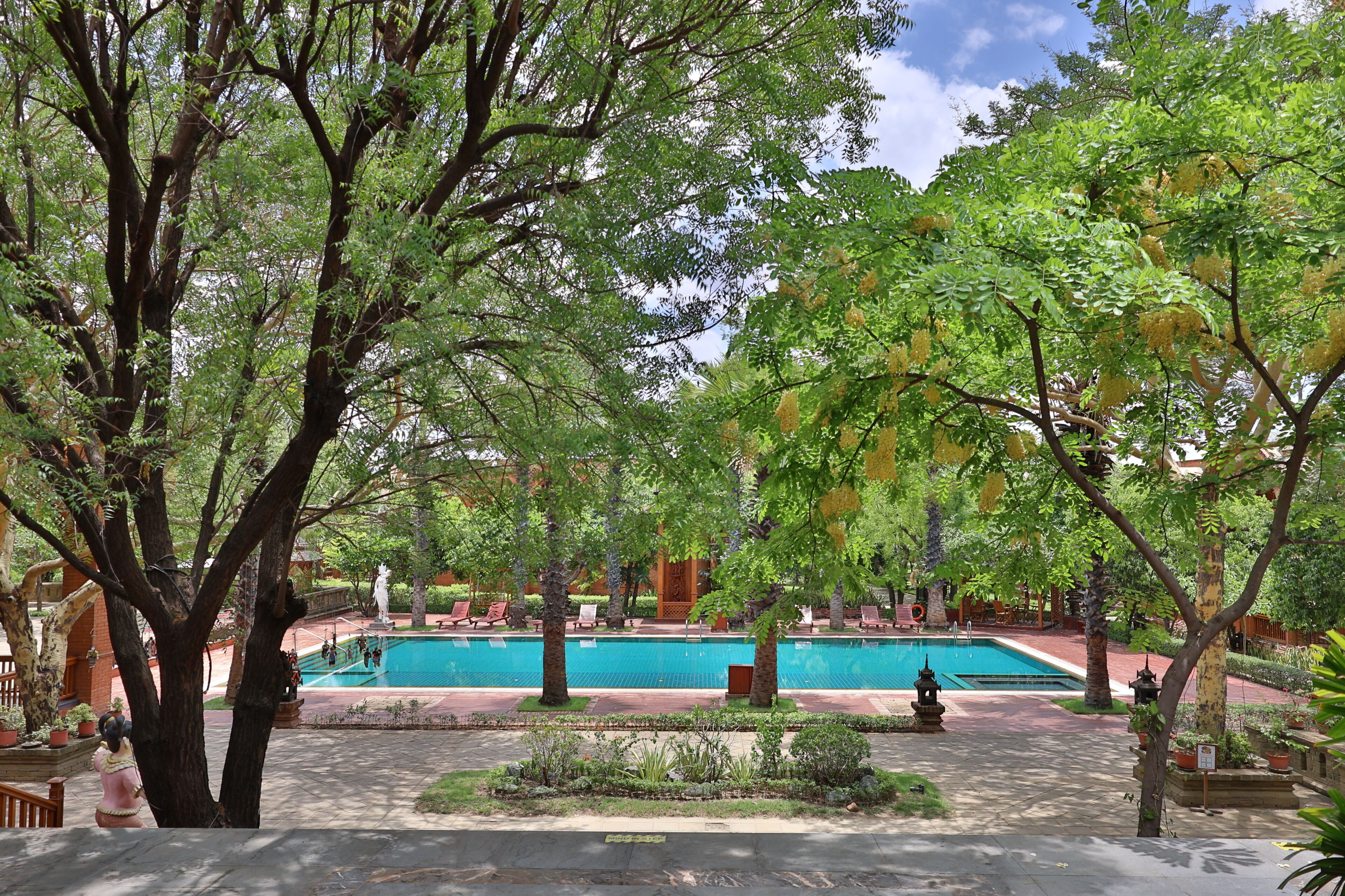 Un área de piscina con una piscina azul cristalina, rodeada de árboles y plantas en macetas.