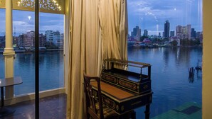 A room with a view of a city skyline across a body of water, a wooden chair, and a table. - Amdaeng Bangkok Riverside Hotel (Bangkok)