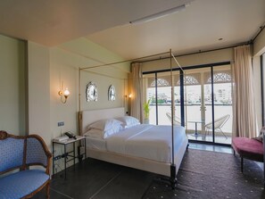 A hotel room with a four-poster bed, a chair, a small table, and a view of the city through large windows. - Amdaeng Bangkok Riverside Hotel (Bangkok)