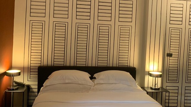 A neatly made bed with white and black bedding, flanked by two bedside tables with lamps, in a room with a patterned wallpaper background. - Amdaeng Bangkok Riverside Hotel (Bangkok)