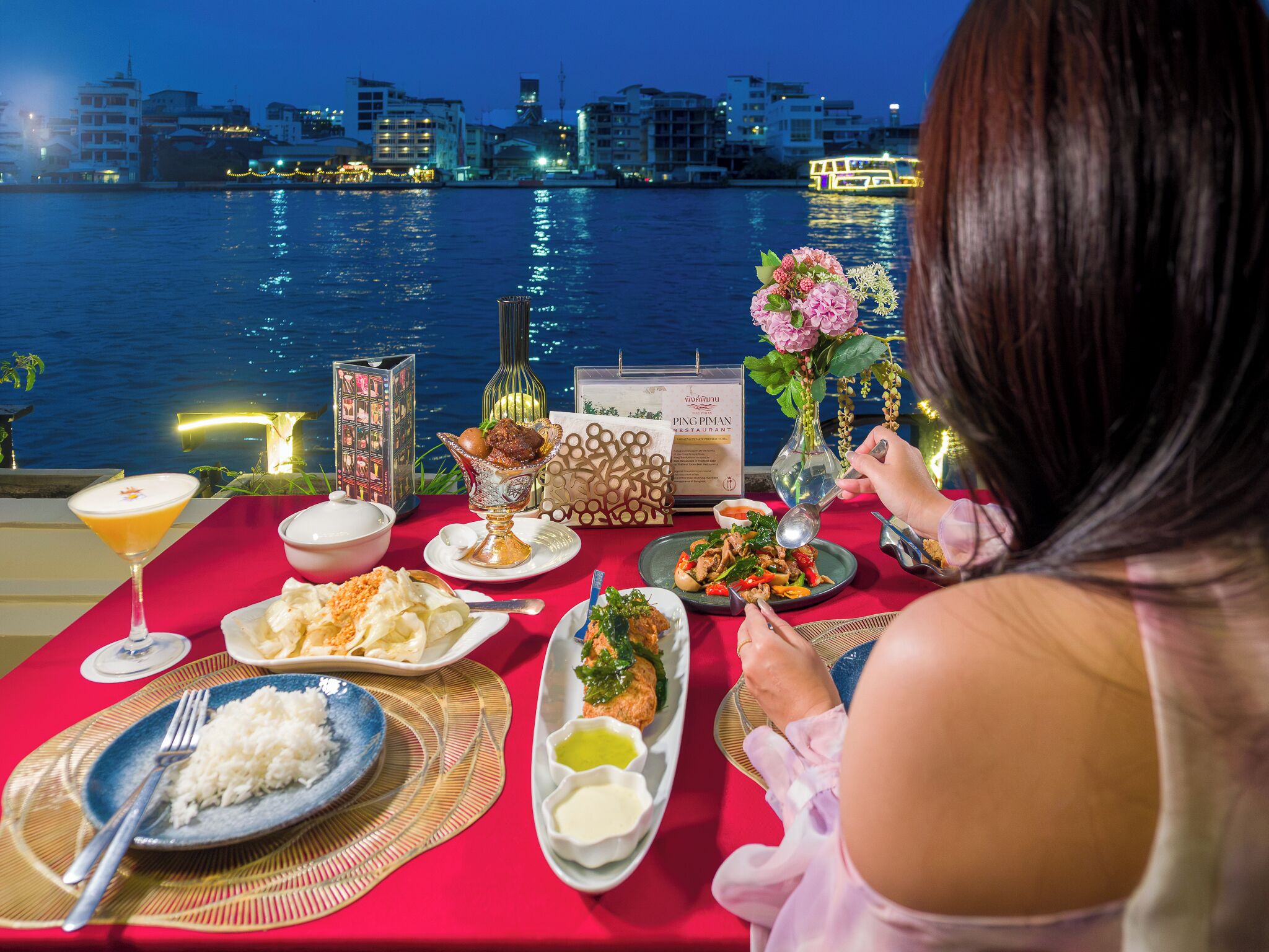 Mesa posta com diversos pratos, um menu e uma garrafa de vinho, com vista para um corpo de água e edifícios ao fundo.