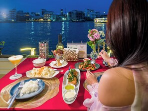 Une table dressée avec différents plats, un menu et une bouteille de vin, donnant sur une étendue d’eau avec des bâtiments en arrière-plan.