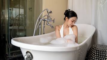 A woman in a bathtub filled with bubbles, with a showerhead and a glass door in the background.