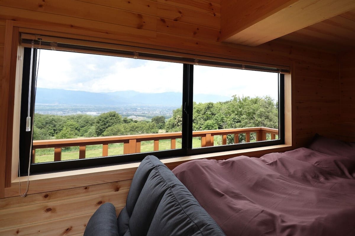 A wooden cabin room with a large window, a bed, and a view of a forest and mountains.