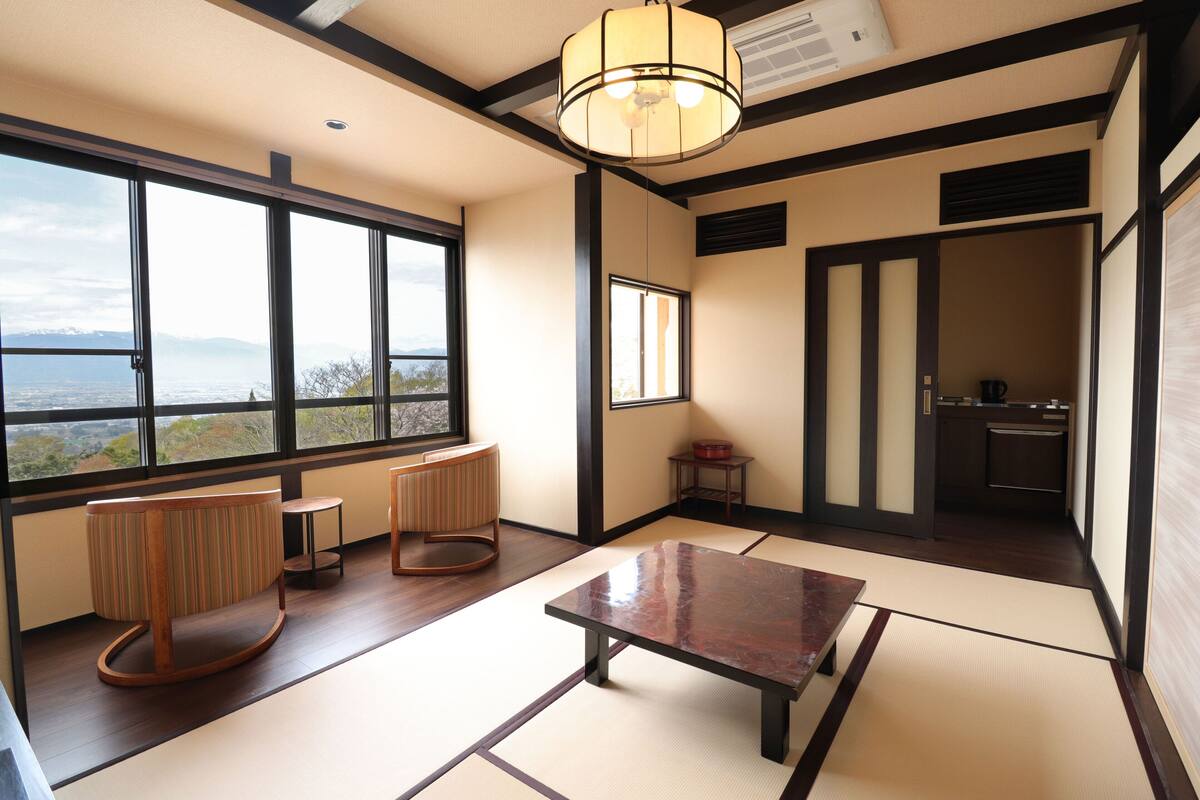 A traditional Japanese room with tatami flooring, a low table, two cushioned seating areas, and large windows offering a view of the mountains.