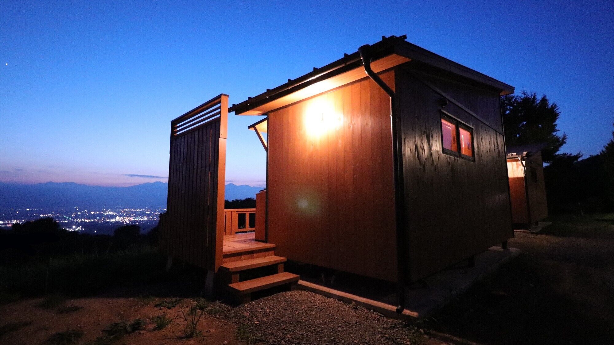 Una pequeña cabaña de madera con una escalera que conduce a una puerta, frente a un cielo crepuscular con luces de la ciudad a lo lejos.