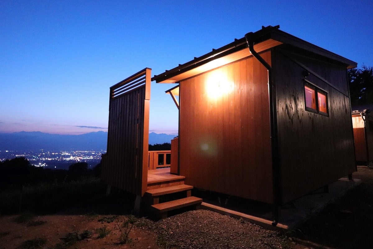 A small wooden cabin with a staircase leading to a door, set against a twilight sky with distant city lights.