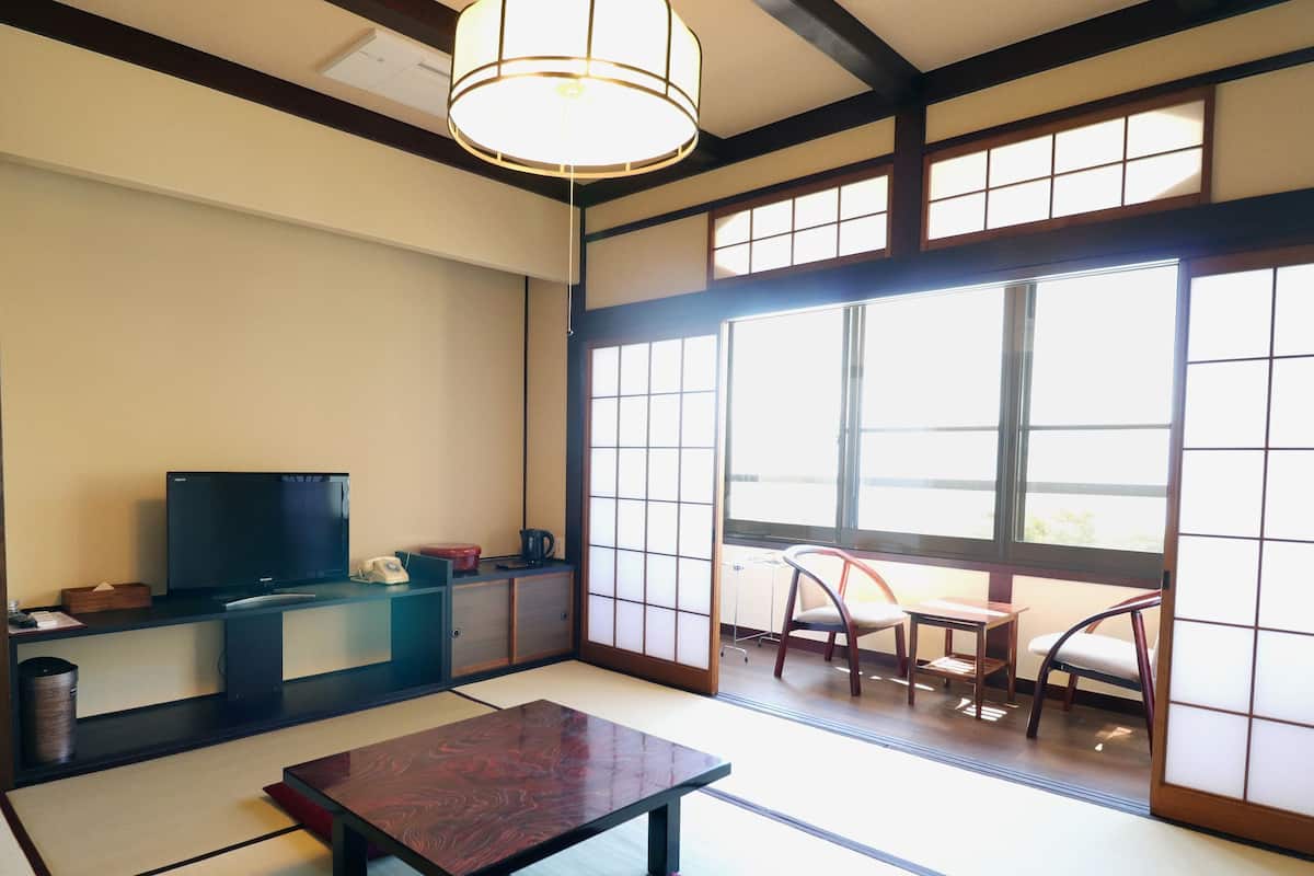 A traditional Japanese room with tatami flooring, a television on a low stand, and sliding doors leading to a balcony with chairs.