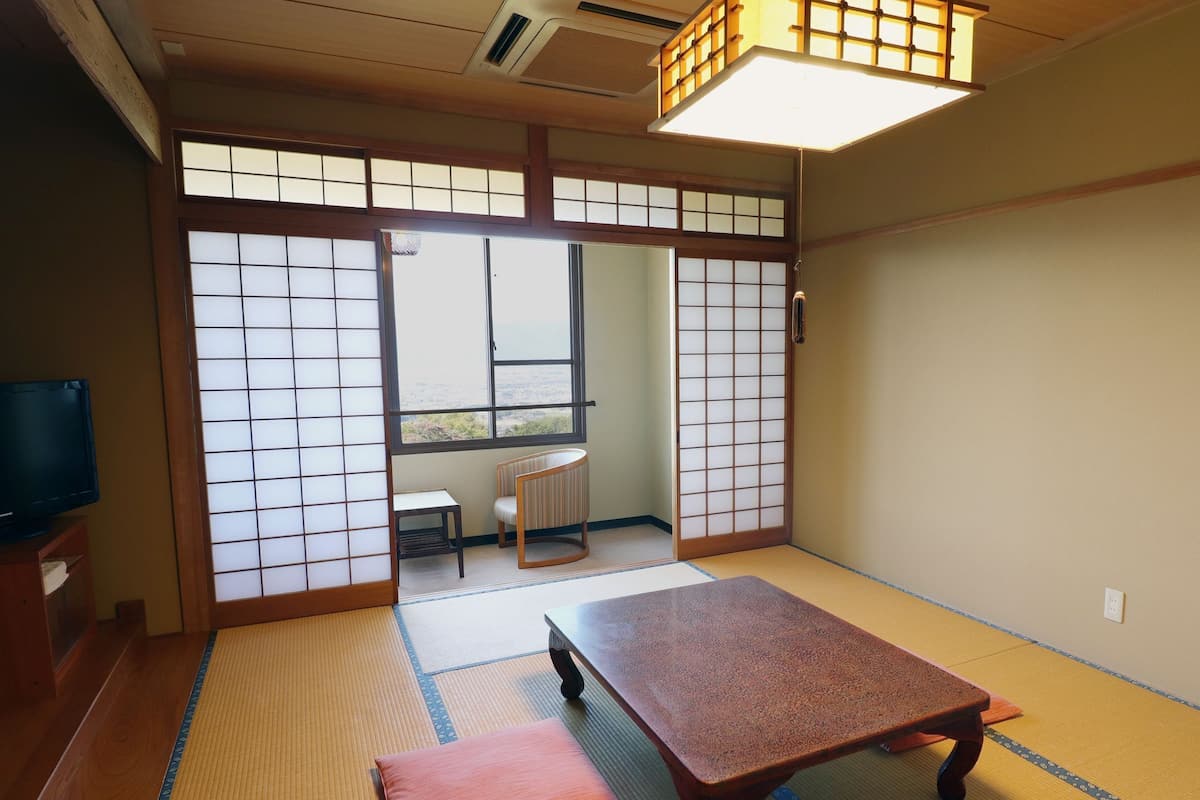 A traditional Japanese room with tatami flooring, a low table, a television on a wooden stand, and sliding doors with a view of the outdoors.