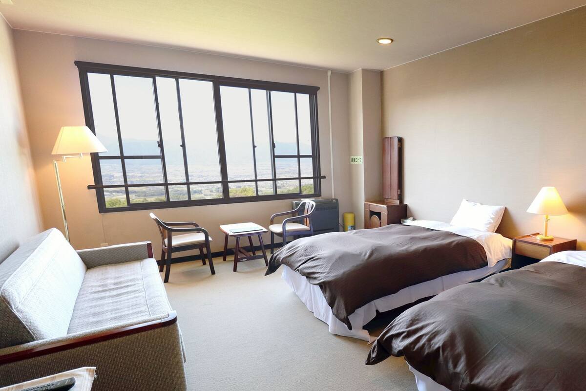 A hotel room with two beds, a desk, a chair, a TV, and a large window with a view.