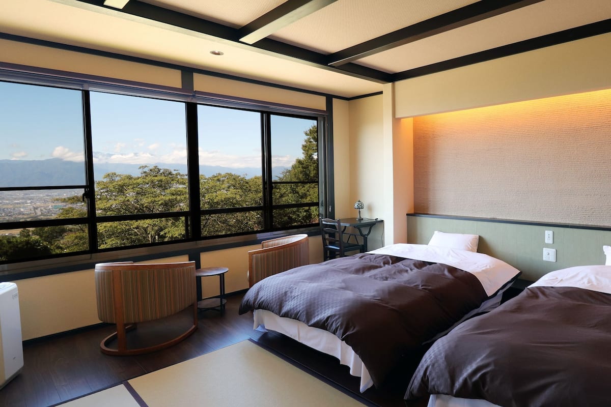 A hotel room with two beds, a view of the city, a small table, and a chair.
