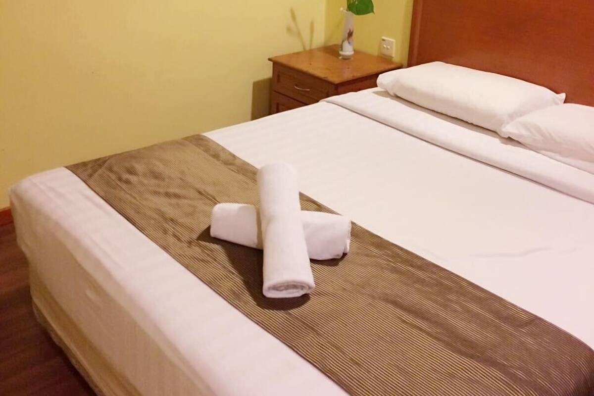A neatly made bed with white linens and a brown accent, a wooden nightstand with a vase of red flowers, and a plain yellow wall.