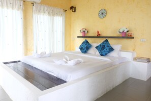 A hotel room with a bed, white curtains, a clock, and a shelf with flowers and figurines.