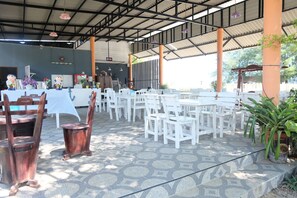Un área de comedor al aire libre con techo, mesas y sillas blancas, piso de baldosas y estructura de techo metálico.