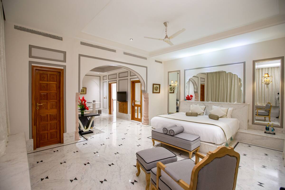 Une chambre d’hôtel avec un grand lit, un coin salon comprenant un fauteuil et un pouf, une télévision et un grand miroir.