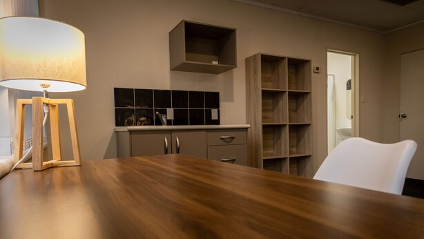 Un coin repas avec une table en bois, une lampe, une chaise blanche et une cuisine en arrière-plan.