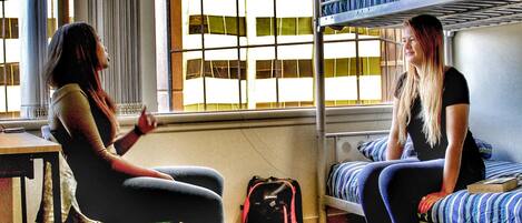 A dormitory room with bunk beds, a wooden floor, and a window with a view of buildings.