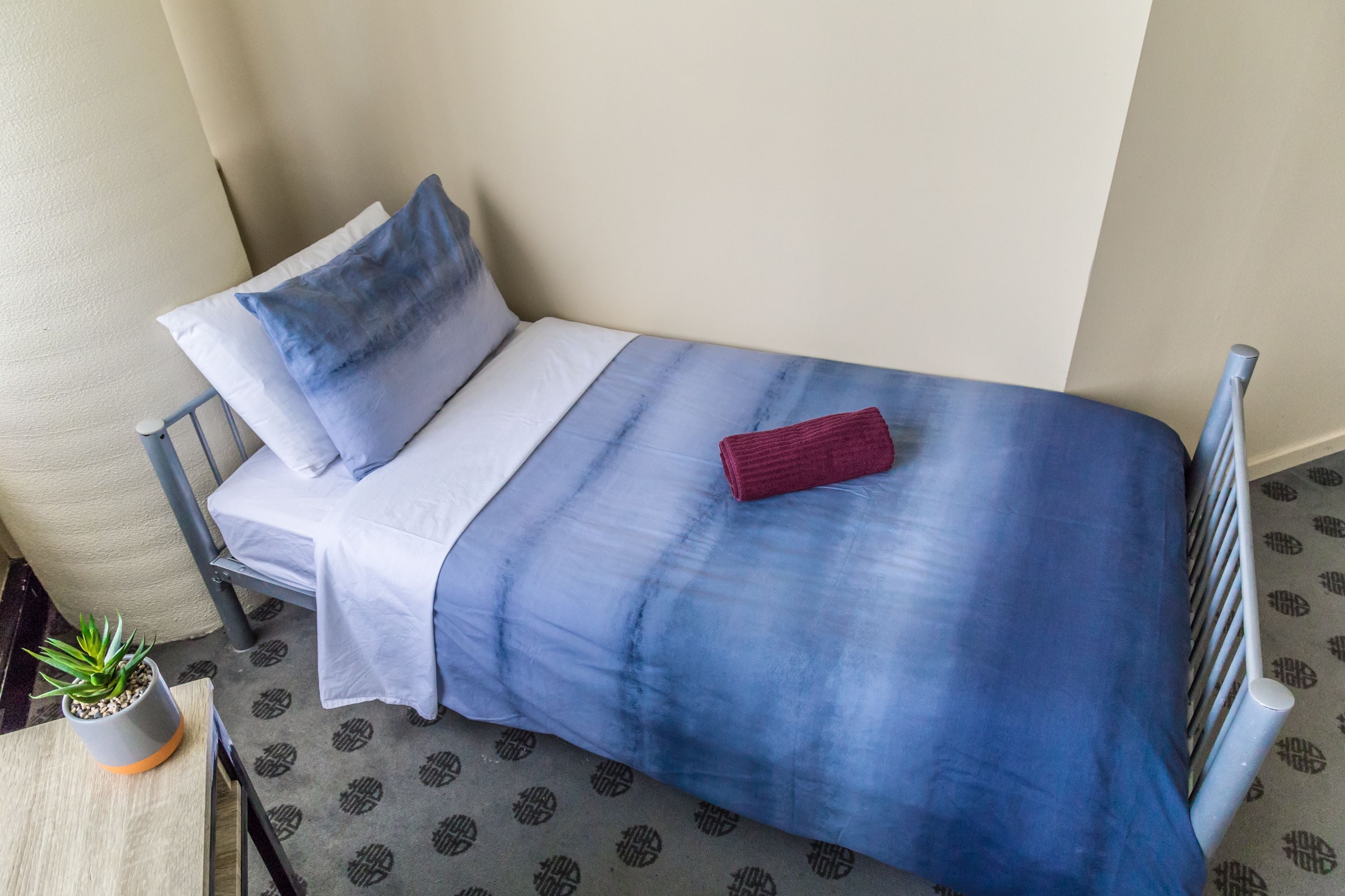Uma cama pequena e bem arrumada, com colcha azul e branca, um travesseiro vermelho e um pequeno vaso de planta ao lado.