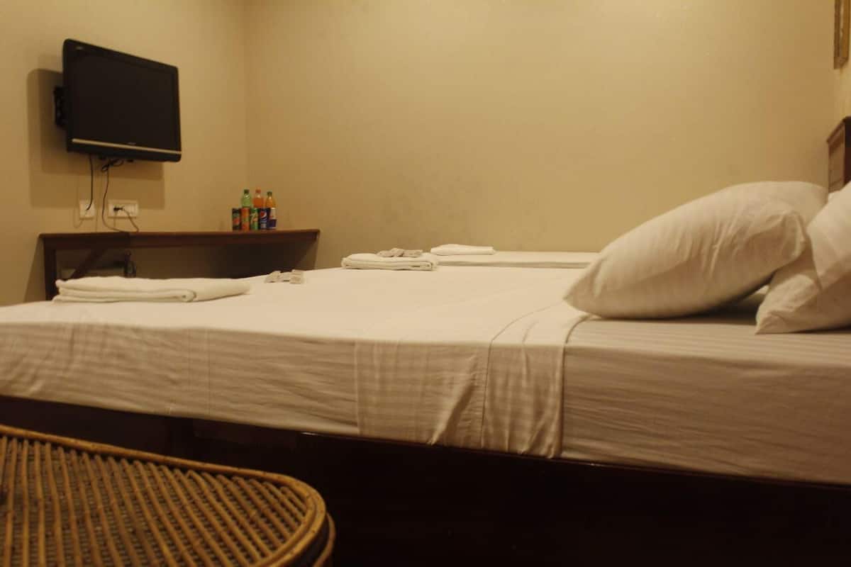 A hotel room with a bed, a television mounted on the wall, a small table with toiletries, and a wicker chair.