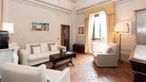 A spacious living room with a white sofa, a smaller armchair, a coffee table, and a fireplace. There are paintings on the walls and a window with curtains. - B&B VillAmorosa (Vicopisano)