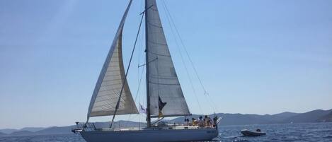 Un velero con varias velas en un mar tranquilo, con una costa lejana y un cielo despejado.